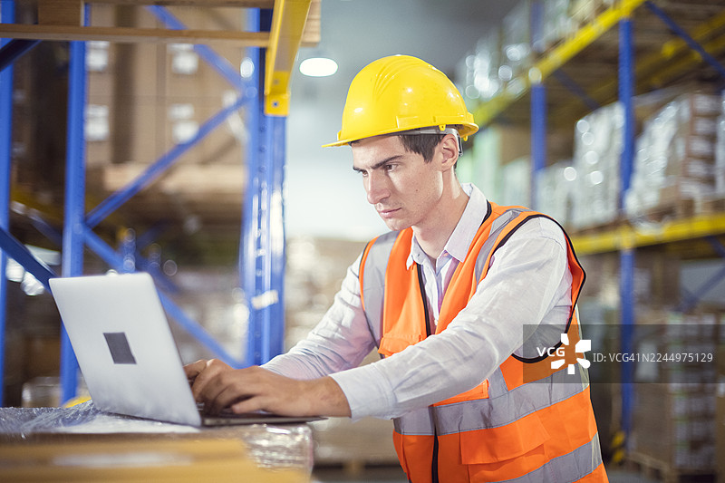 仓库工人戴着安全帽和反光背心，使用笔记本电脑优化库存管理，提高物流效率。图片素材