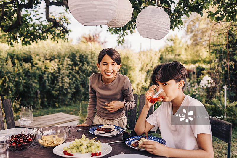 花园派对上，微笑着的女孩站在喝果汁的哥哥旁边图片素材