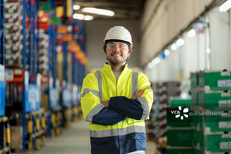 一位面带微笑的男性物流工程师身着安全装备，自信地站在宽敞的仓库中，双臂交叉，代表着技术驱动的库存管理、团队合作以及供应链、物流和现代工业运营的效率。图片素材