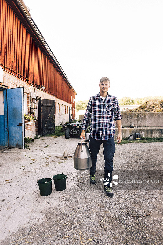 一名牧牛工人手持奶桶，在谷仓前行走的全景照图片素材