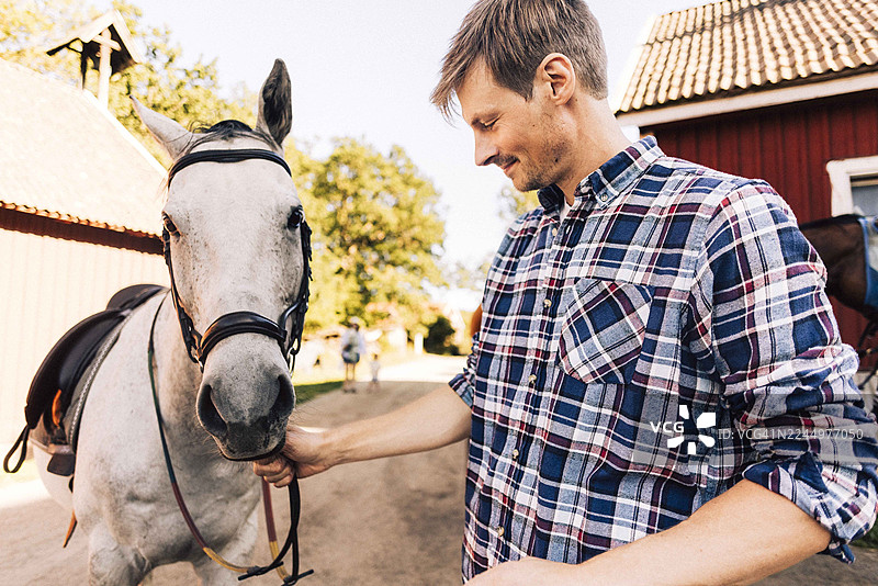 穿着格子衬衫、面带微笑的农夫在谷仓旁牵着马的缰绳图片素材