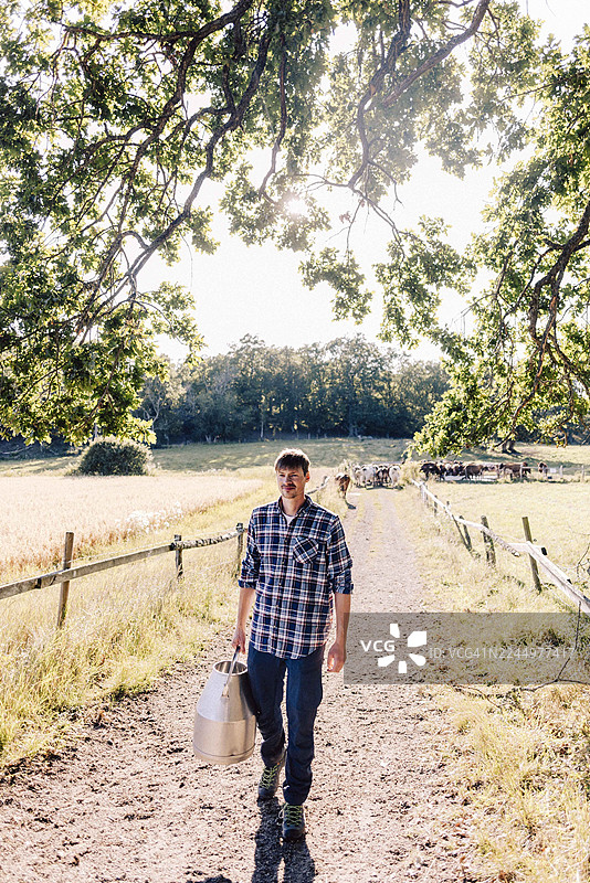 一位农民走在土路上，手持奶罐，神情充实。图片素材