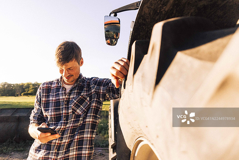 农场里，一位农民微笑着靠在拖拉机轮胎上，同时正在使用智能手机。图片素材