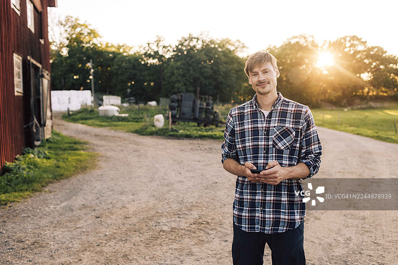 一位面带微笑的男性农民站在农场里，手中拿着智能手机。图片素材