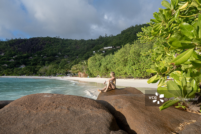 女子坐在大岩石上，欣赏着沙滩和热带海洋的景色，地点为塞舌尔马埃岛的佩蒂特安塞海滩图片素材