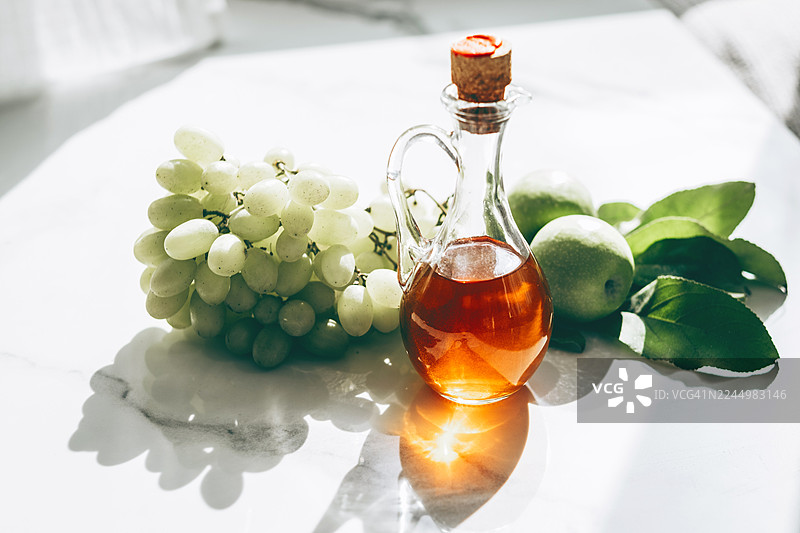 白色葡萄、带叶子的青苹果和一瓶软木塞封口的金黄色苹果醋，在柔和的自然光下陈列在大理石台面上。图片素材