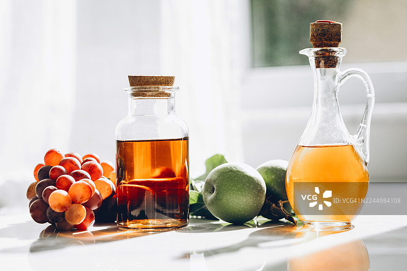 浅色大理石台面上摆放着装有苹果醋和葡萄醋的玻璃瓶，旁边散落着红葡萄、青苹果和新鲜叶子。光线明亮自然，阴影清晰。图片素材