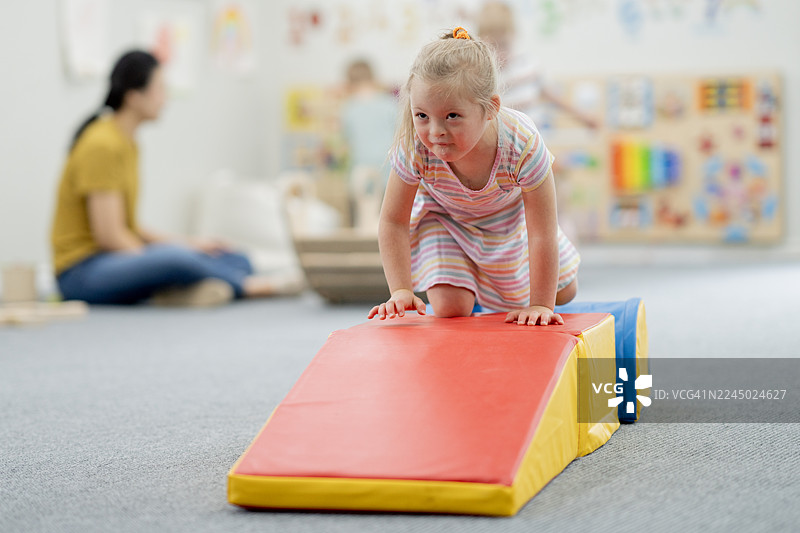保育员在旁，小女孩在日托游戏室的彩色软坡上，专注而好奇图片素材
