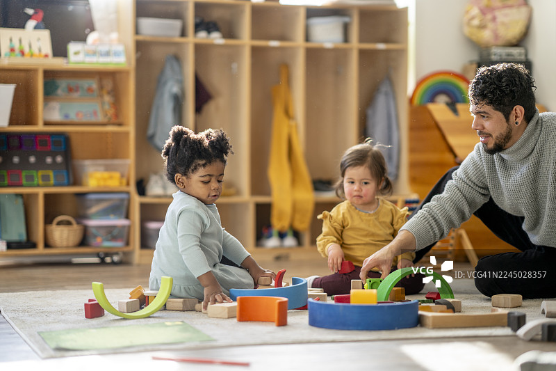 多元化的日托场景，一位充满关爱的成年人在明亮的教室里指导两个孩子玩耍图片素材