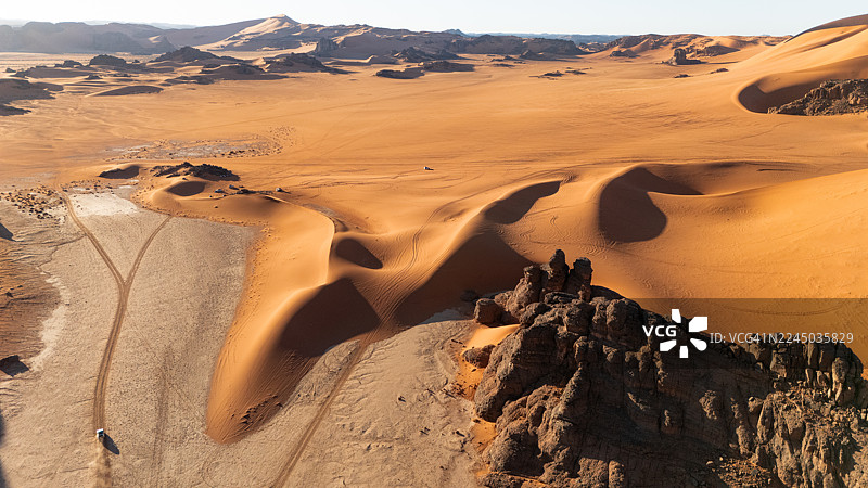 蔚蓝天空下，广阔起伏的沙漠景观令人惊叹的航拍景象，展示了沙丘、岩石构造和轮胎痕迹，位于阿尔及利亚贾内特，塔德拉尔特。图片素材