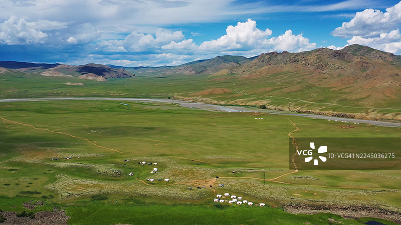 从空中俯瞰蒙古广阔的绿色山谷，一条蜿蜒的河流、传统的蒙古包和远处的山脉在多云的天空下若隐若现。图片素材