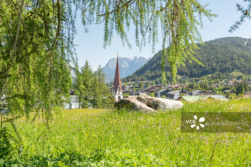 阳光明媚的日子里，风景如画的阿尔卑斯山村，一座醒目的教堂尖顶坐落在一片郁郁葱葱的绿色山谷中，周围环绕着雄伟的山脉和森林，蒂罗尔的泽费尔德，奥地利图片素材