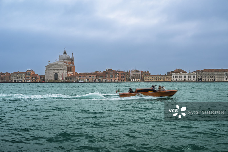 一辆水上出租车在威尼斯泻湖上飞驰，背景是朱代卡岛上历史悠久的建筑和救世主教堂，天空多云，地点是意大利威尼斯。图片素材