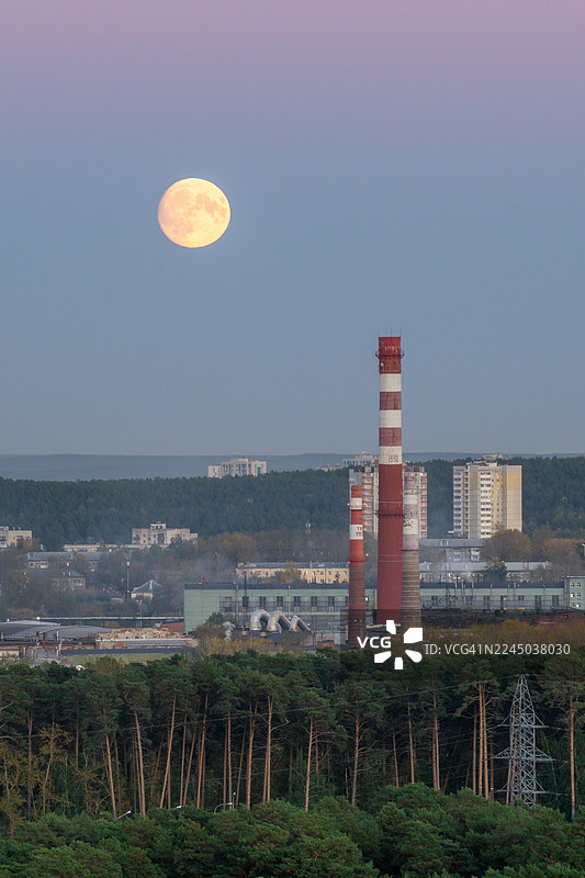 暮色中，满月升起在工业景观之上，高耸的烟囱和前景中茂密的森林图片素材