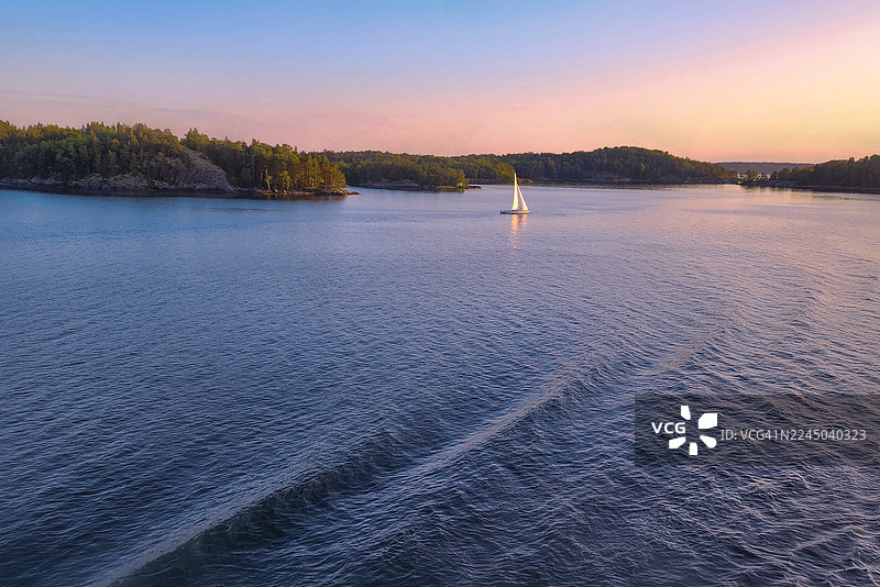 宁静的日落帆船：平静水面上森林岛屿背景下的帆船美景，瑞典，瓦克斯霍尔姆图片素材