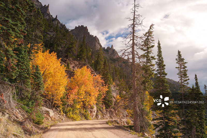 积雪覆盖的山脉，秋日色彩斑斓的树木和常绿森林，在多云的天空下蜿蜒穿过一条土路，位于美国埃斯特斯公园。图片素材