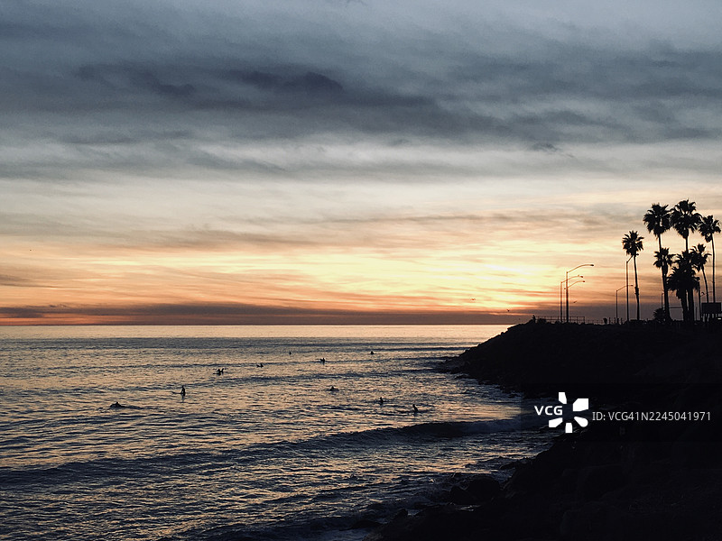 日落时分，海景与戏剧性的天空交相辉映，海岸线上剪影般地映着冲浪者和棕榈树。图片素材