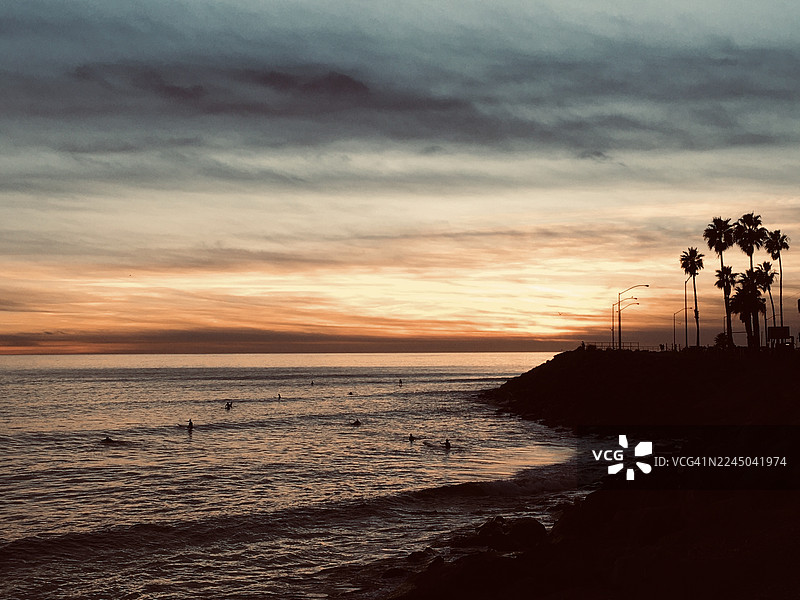 日落时分，壮丽的天空映衬着海景，岸边是冲浪者和棕榈树的剪影图片素材