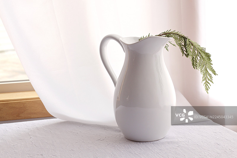 窗边，薄纱窗帘旁，纹理桌上摆放着一个优雅的白色陶瓷水壶，旁边有一枝绿叶，营造出宁静简约的意境。图片素材