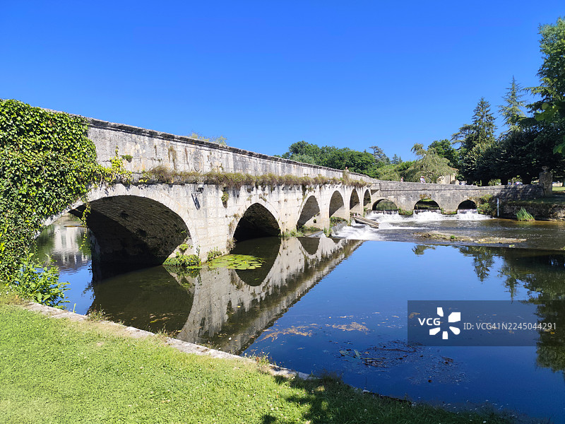 一座历史悠久的石头拱桥横跨宁静的河流，天空湛蓝，四周绿树成荫，位于法国。图片素材