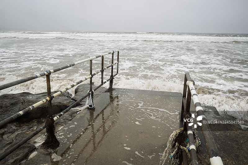 阴天时，汹涌的海浪拍打着带有金属栏杆的混凝土码头，构成了一幅戏剧性的海岸景象图片素材