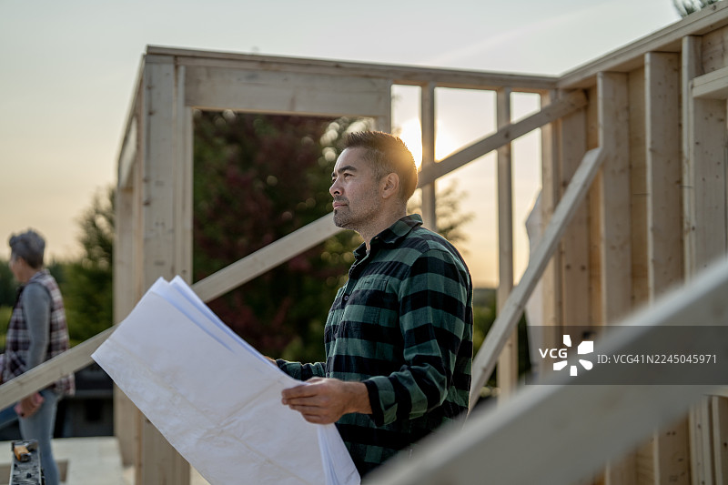 日落时分，一名亚洲男性建筑工人正在户外建筑工地分析蓝图，旁边有计划和草稿。图片素材