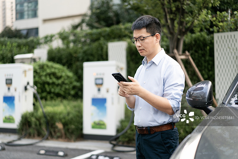 城市中，人们在电动汽车充电站使用智能手机，背景有汽车和建筑图片素材