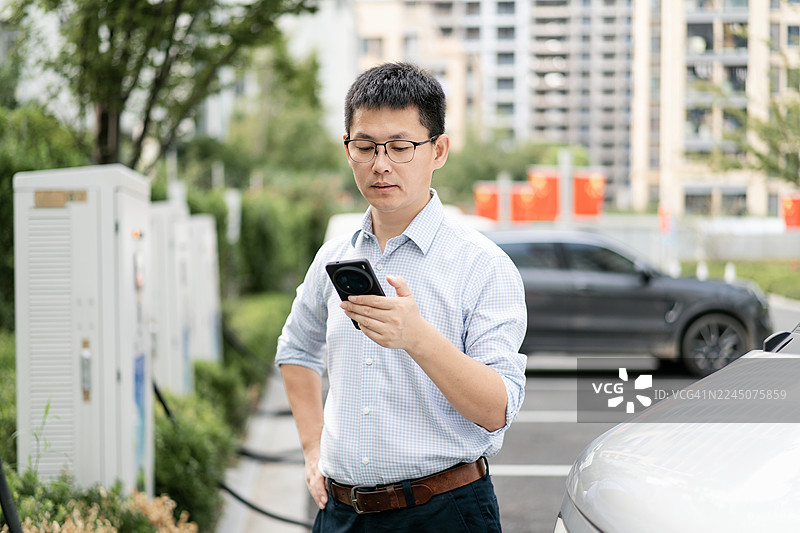 在城市中，人们在电动汽车充电站使用智能手机，背景有汽车和建筑物。图片素材