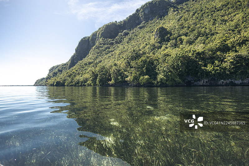宁静湖泊清澈的水面倒映着郁郁葱葱的绿色山景，天空湛蓝。印度尼西亚马鲁古省塞尔姆岛塞尔曼村。图片素材