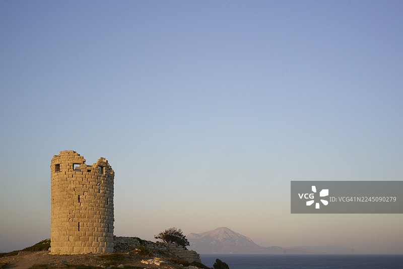 希腊伊卡里亚岛的古希腊德拉科农塔楼。古老的石塔在日落时分，在晴朗的蓝天下俯瞰着宁静的大海。图片素材