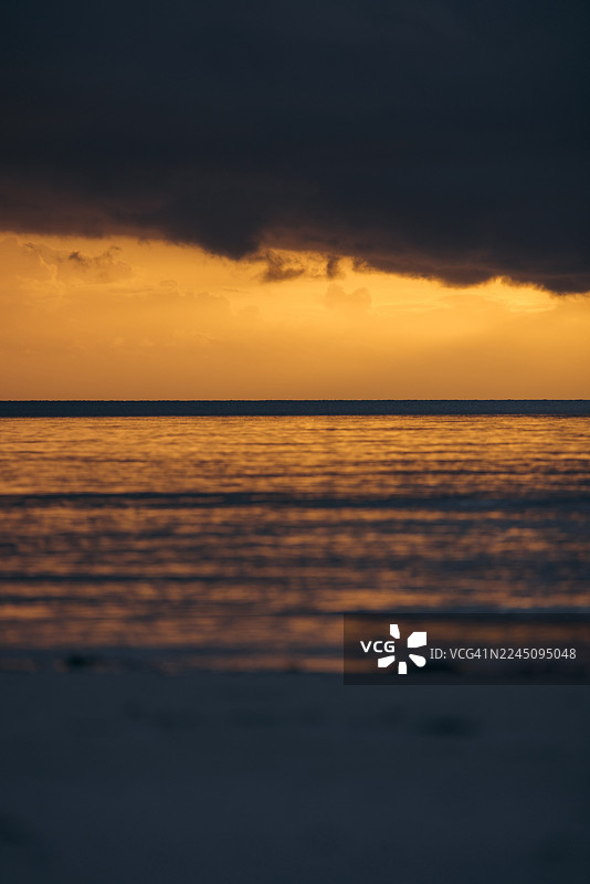 深色云彩笼罩着宁静的日落海洋，温暖的橙色光芒倒映在平静的海面上。印度尼西亚东南马鲁古省凯奇尔岛的恩格布洛海滩（Pantai Ngurbloat or Ngurbloat Beach）。图片素材