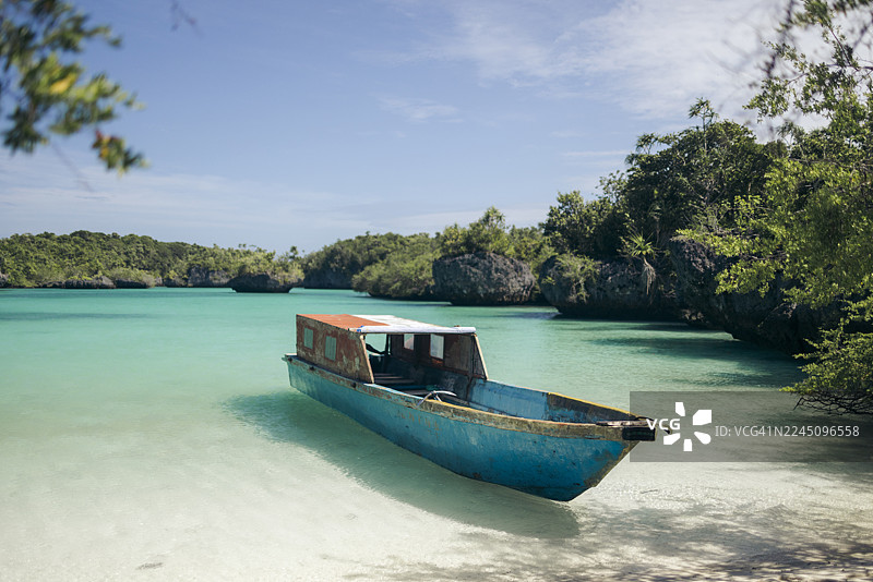 阳光明媚下，一艘蓝色小船停靠在绿意盎然的热带海岸边，水面呈现出绿松石般的色彩。摄于印度尼西亚马鲁古省东南马鲁古县的凯奇尔岛（Pulau Bair / Bair Island）。图片素材