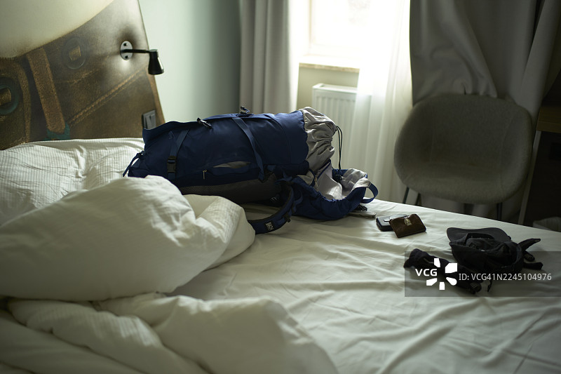一个蓝色的徒步背包和衣服散落在舒适、光线柔和房间的白色床上。德国慕尼黑。图片素材