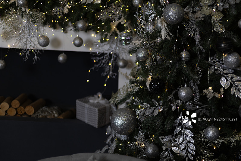 一棵装饰华丽的圣诞树，挂着银色装饰球、闪光叶子和彩灯，矗立在深色壁炉架旁，旁边有一个银色礼品盒和一堆柴火。图片素材