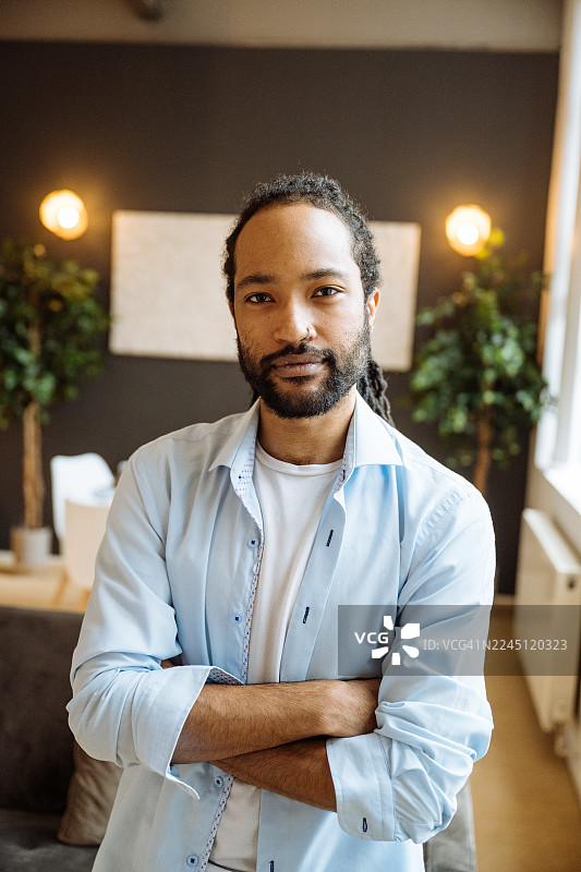 留着胡须的黑人男子面带微笑，坐在深色沙发上，身旁环绕着室内植物，柔和的光线照射着他，他正看向镜头。图片素材