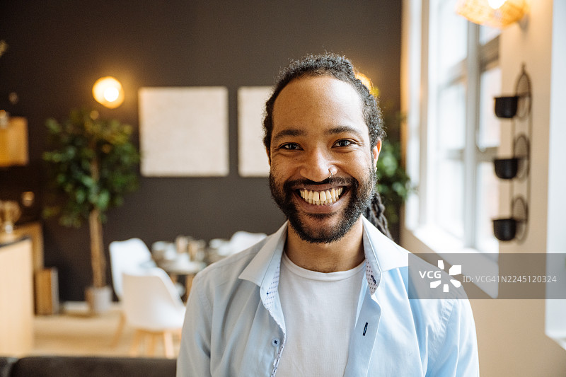 一个留着胡须的黑人男子面带微笑，坐在深色沙发上，周围环绕着室内植物和柔和的光线，正看向镜头。图片素材