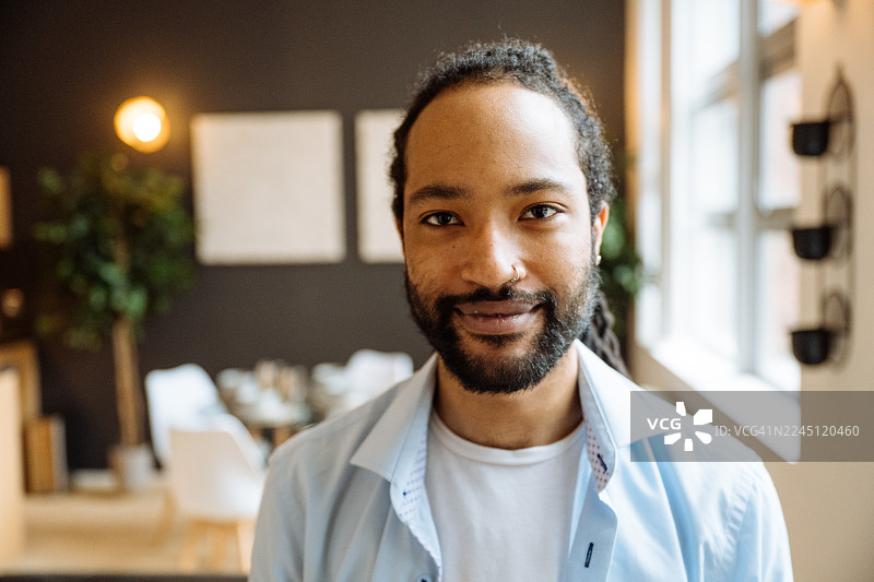 留着胡子的黑人男子面带微笑，坐在深色沙发上，周围环绕着室内植物和柔和的灯光，他正看向镜头。图片素材