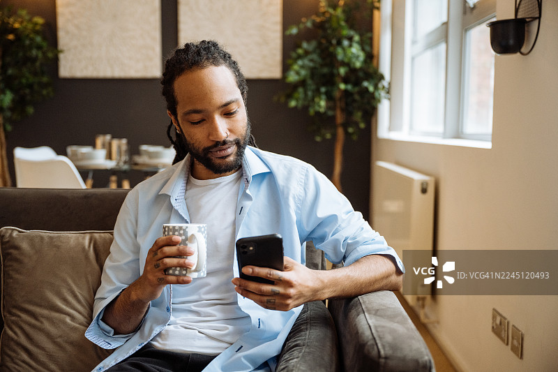 留着胡子的黑人男子面带微笑，坐在深色沙发上，周围环绕着室内植物和柔和的灯光，他正在使用智能手机。图片素材
