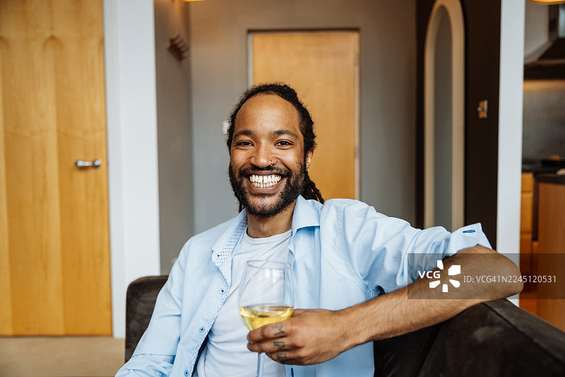 留着胡子的黑人男子微笑着坐在深色沙发上，周围环绕着室内植物，他手里拿着一杯白葡萄酒。图片素材