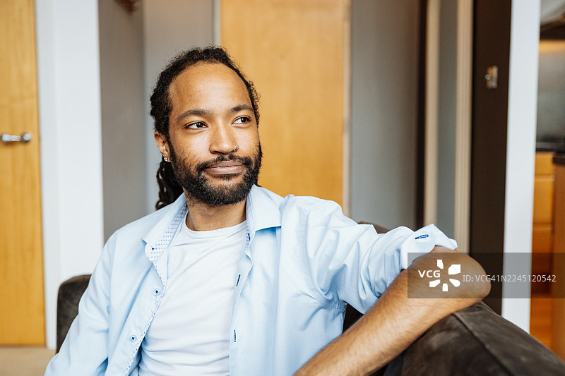 留着胡须的黑人男子面带微笑，坐在深色沙发上，周围环绕着室内植物和柔和的光线图片素材