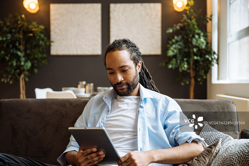 留着胡须的黑人男子面带微笑，坐在深色沙发上，周围环绕着室内植物，他正使用平板电脑。图片素材