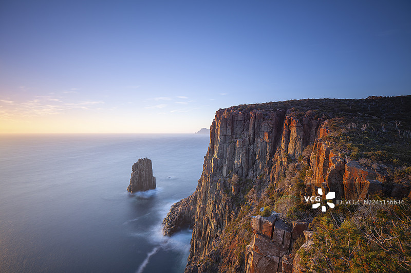 日出时的长曝光照片，展示了豪伊角（Cape Hauy）壮丽悬崖上发光的黎明。望向 the Cape Pillar。温柔的海浪拍打着陡峭的悬崖。澳大利亚塔斯马尼亚塔斯曼半岛的豪伊角。图片素材