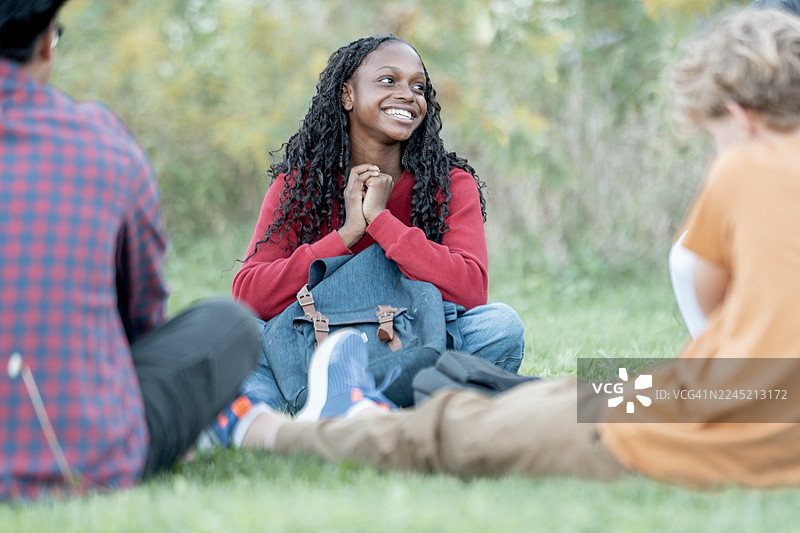 一位穿着红色毛衣、背着背包的微笑黑人女性，在户外公园友好地与人交谈图片素材