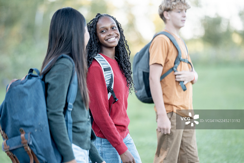 一群来自不同背景的微笑学生，背着背包在绿意盎然的公园里行走图片素材