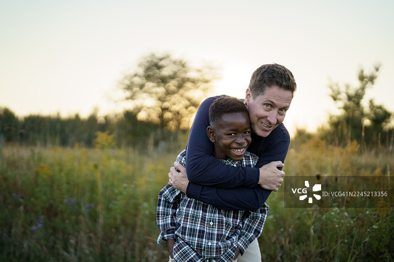 不同种族父子在夕阳下的田野里拥抱，庆祝家庭与友谊图片素材