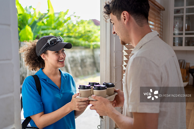 一位身穿蓝色制服的拉丁裔送餐女郎在家中为顾客送饮品。一位迷人的女性快递员在前门拿着外卖咖啡杯，象征着食品配送和在线订购服务。图片素材