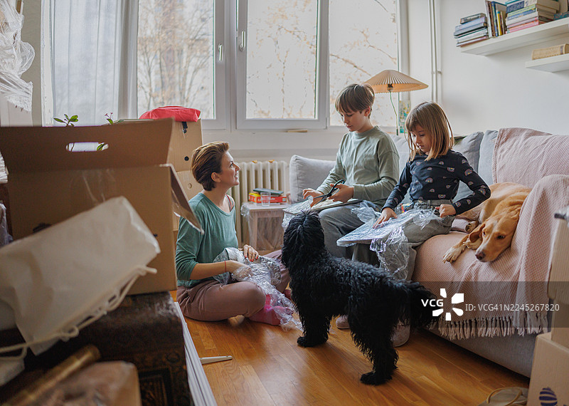 女人坐在地板上，旁边是狗，孩子们坐在沙发上，房间里堆满了箱子。图片素材