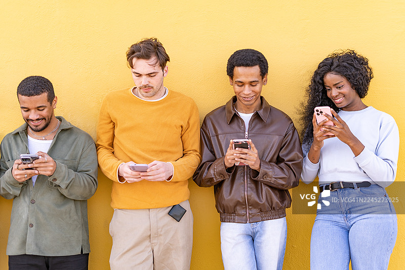 四个朋友站立在黄色墙壁前，各自看着手机，并望向镜头图片素材
