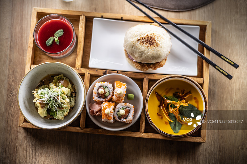 这张照片展示了一个装满各种日本美食的便当盒。寿司卷、迷你包子汉堡、红薯汤、西兰花天妇罗和奇亚籽布丁等美食被分别盛放在木托盘上的碗碟里。图片素材