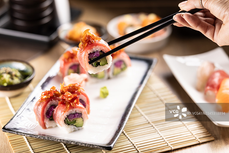 这张照片捕捉到一只手用筷子夹着一块金枪鱼包裹的乌拉玛基寿司卷，上面有焦糖洋葱，正悬在盘子上方。图片素材